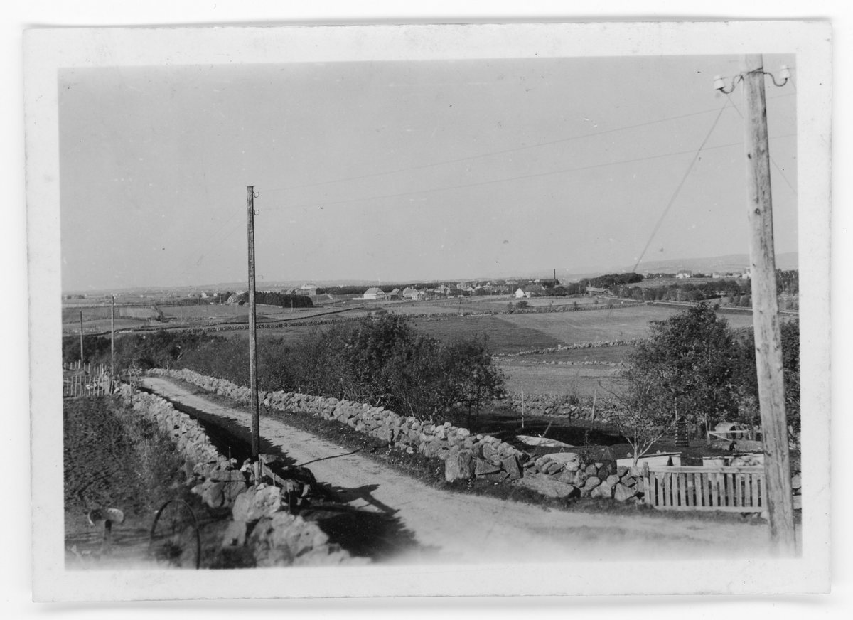 Bildet viser Kvia. Eit jordbrukslandskap. I forgrunnen går ein grusveg med steingard og telefonstolpar på begge sidene. Busetnad i bakgrunnen.