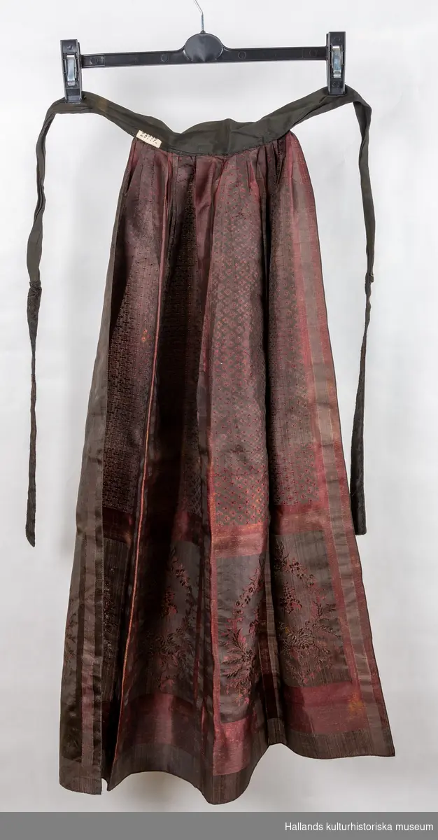 Förkläde, brudförkläde av siden. Mönstrat i rödbruna färger. Mått: bredd upptill 20 cm, bredd nedtill 75 cm.