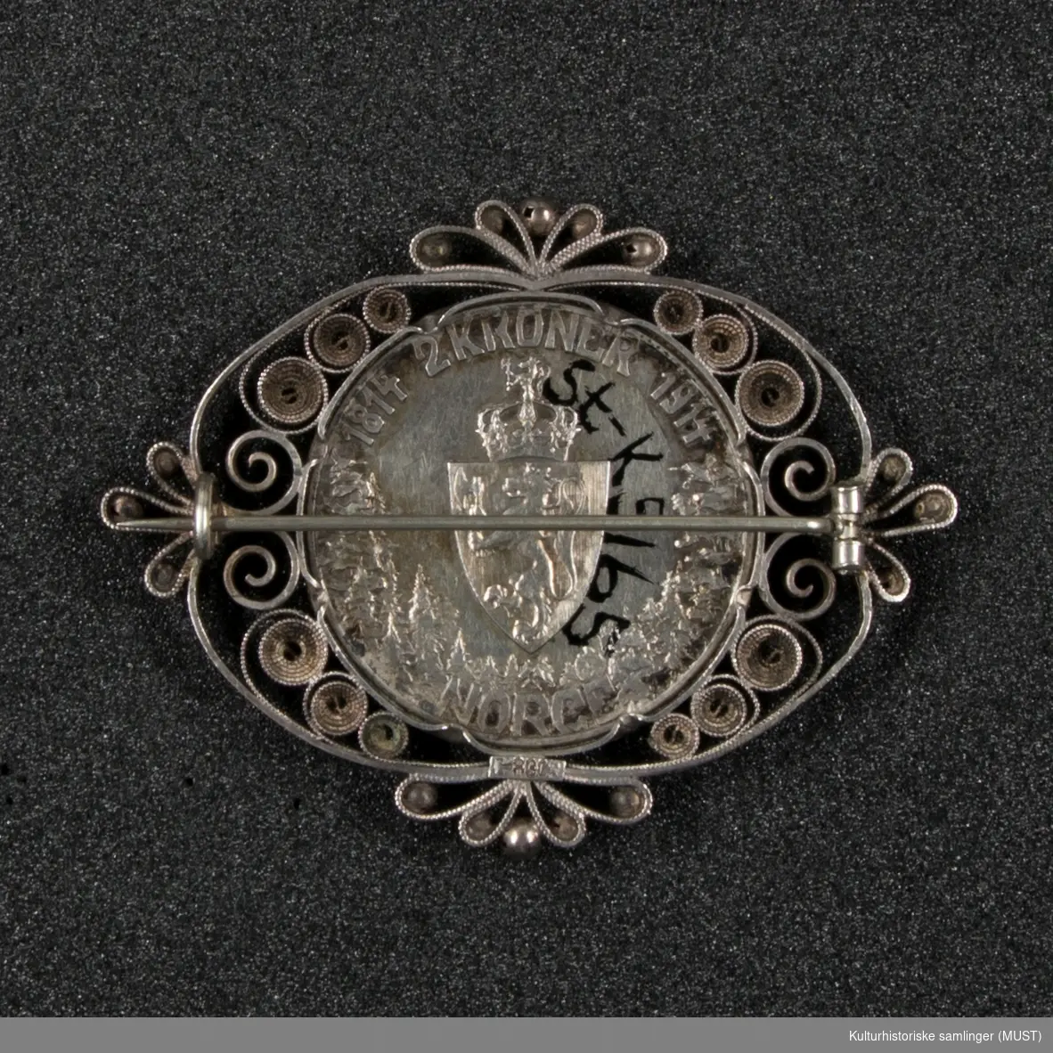 Brosje laget av en 2 kroning fra 1914. Myntens har på den ene siden Norges våpenskjold; løve med krone og på den andre siden vises en kvinne med kjole. Filigrans-dekor rundt mynten.