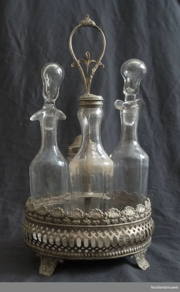 Stativ med fire glassflasker/beholdere som settes på matbordet. Metallstativet er utformet som en liten kurv med lang hank, alt med floral dekorasjon.