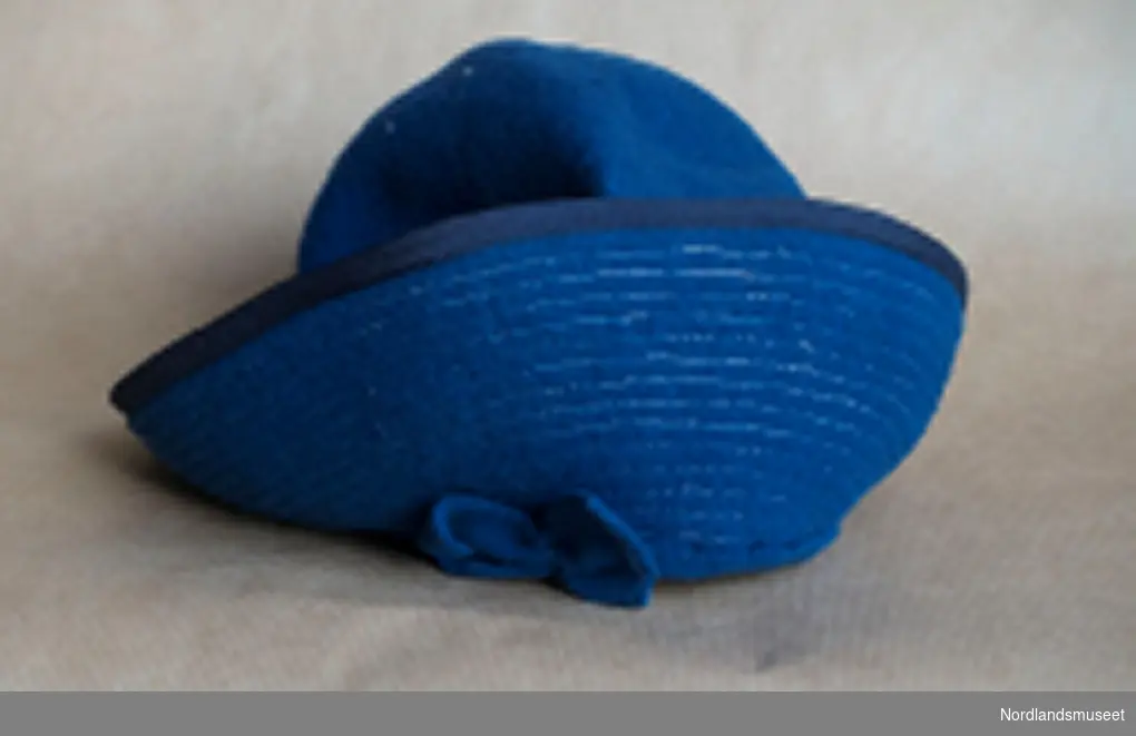 Liten blå filthatt, damehatt eller jentehatt, opprettet kant med blå filtsløyfe foran og bak. Tynt gummibånd til å feste under haken.