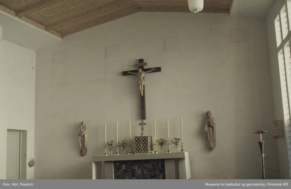 Bildet viser interiøret til Den katolske kirke St. Mikael i Hammerfest. I midten ser vi alteret som er pyntet med duk, stearinlys og blomsterbuketter. På alteret står tabernakelet som tjener til oppbevaring av hostiene. Ovenfor henger det en fremstilling av Jesus på korset, skjært i tre. Ved siden av alteret er det to ytterligere treskulpturer på veggen.