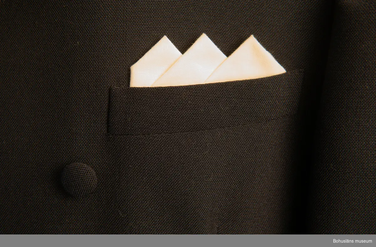 Marinblå kostym med dublla kanpprader och vit duk i vänster bröstficka. Kostymen måttsydd till givarens bröllop. Fodrad med paisley mönstrat tyg. Samma mönster på slips UM033940.

En sprund på baksida.