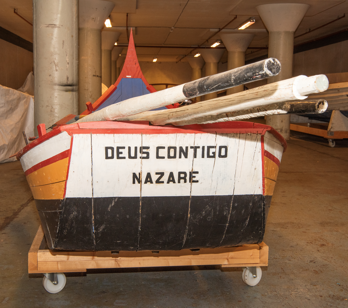 Liten portugisisk robåt med flat bunn, spiss og høy baug. Innvendig sort bunn, blå tiljer/tofter, rød esing mm. Utv. sort bunn, oker, hvit med røde streker, blå.