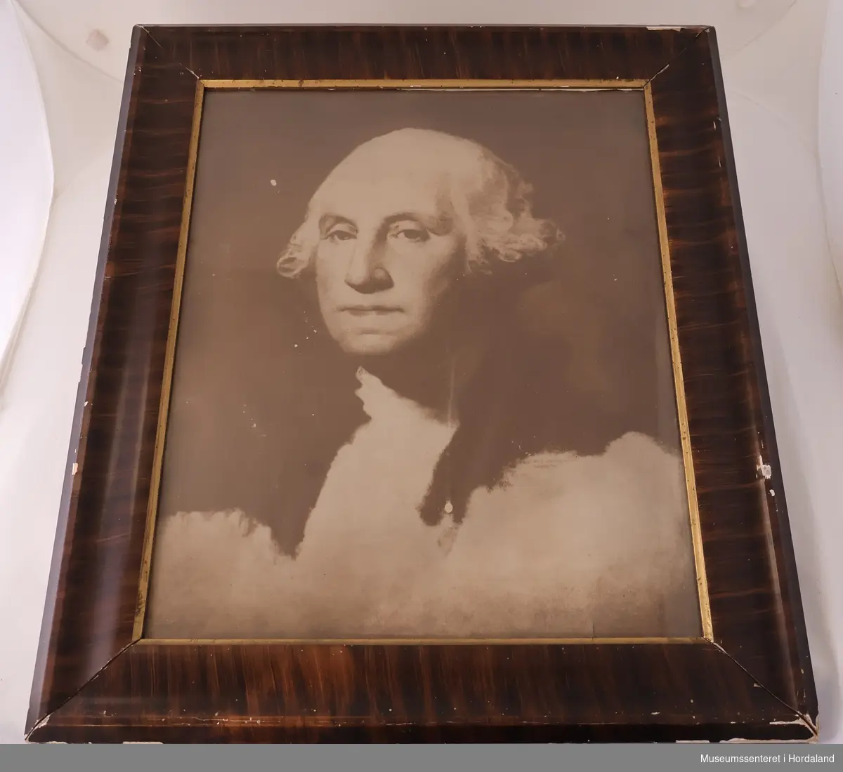 Portrett av George Washington på papir i sepiatoner, bak glass og i rektangulær, profilert treramme med en indre forgylt kant.