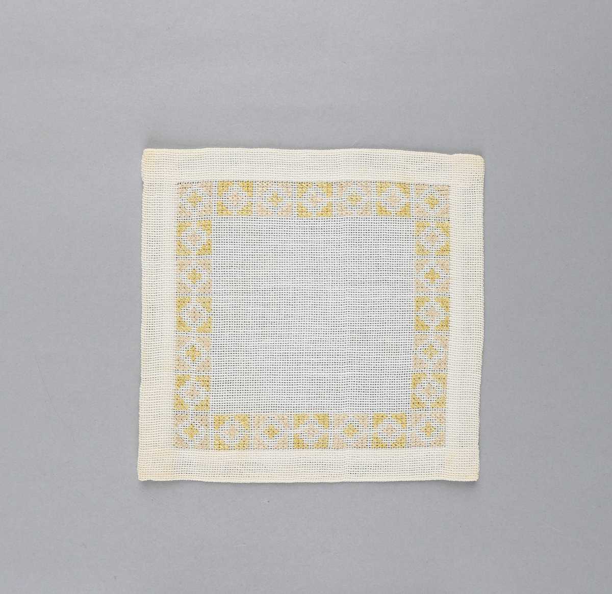 Tilnærmet kvadratisk dekkebrikke i hvitt tekstil med brodert bord langs brettekanten. Broderiet består av korssting av gul og rosa tråd i geometrisk mønster.
