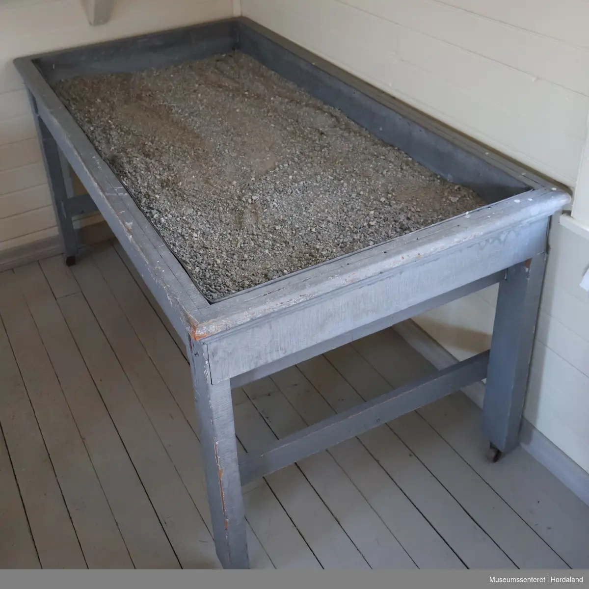 Rektangulært, gråmalt bord på fire bein med hjul. Bordplaten er en sandkasse fôret med sink. Oppi sinkbaljen er det grov sand.