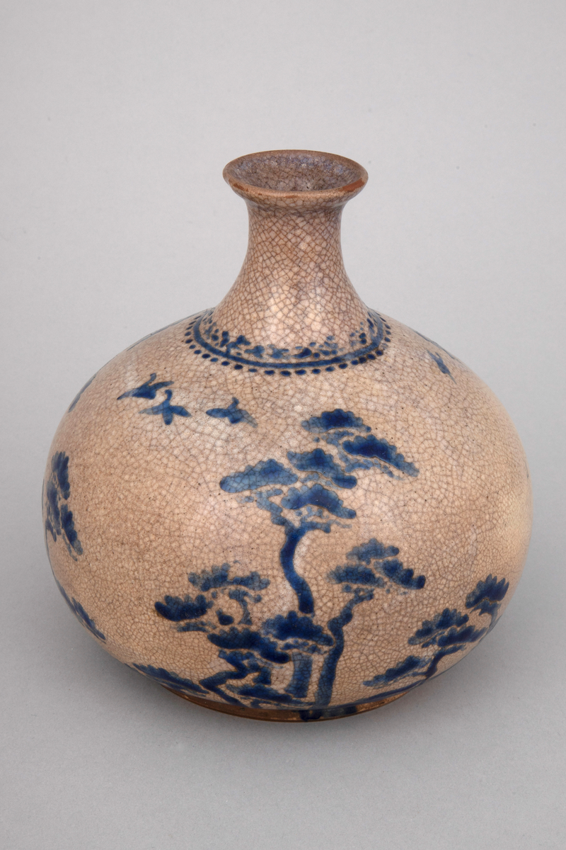 Vase i dreiet keramikk. Gråbrun glasur med krakeleringer. Dekor i blå overglasurmaling. Glasuren dekker ikke fotrandens underside. Dekoren viser motiv med stiliserte tre.