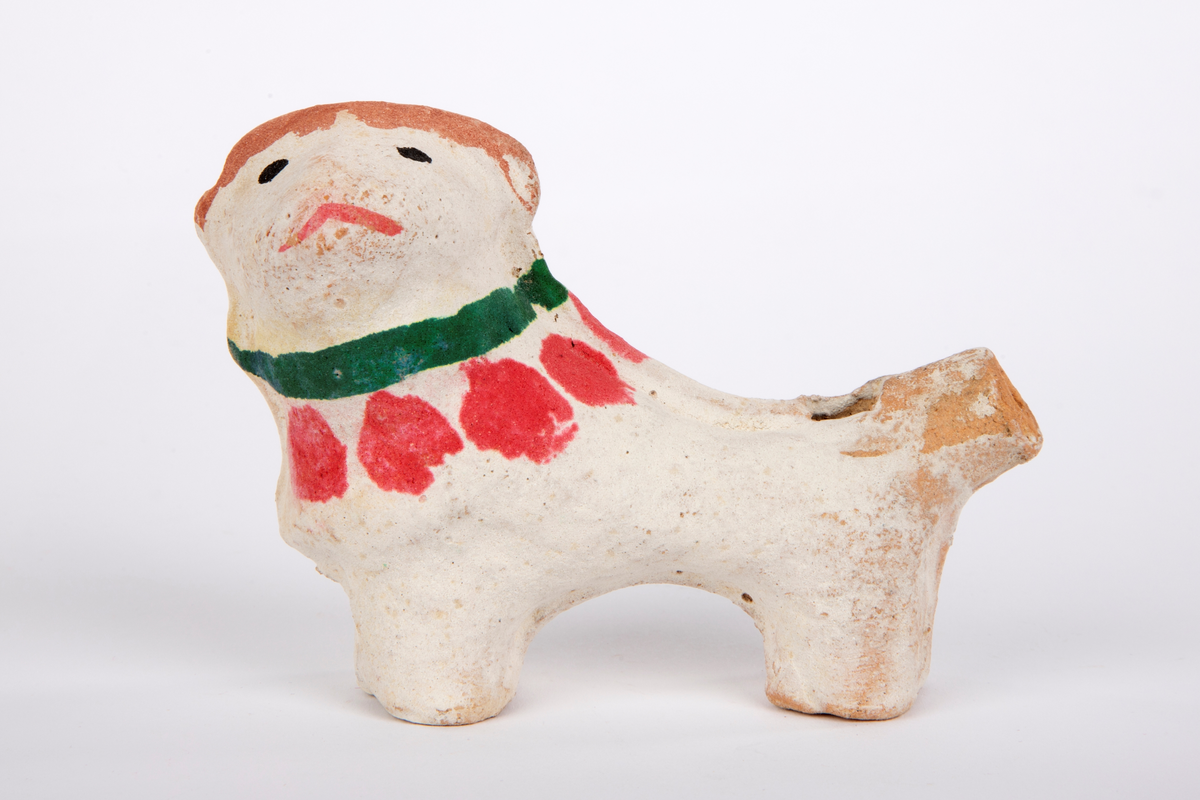 Fløyte i leire, formet som en slank, hvitmalt hund med brun stripe på hode og ører. Svarte øyne og rød munn, grønt halsbånd og røde flekker rundt halsen.
