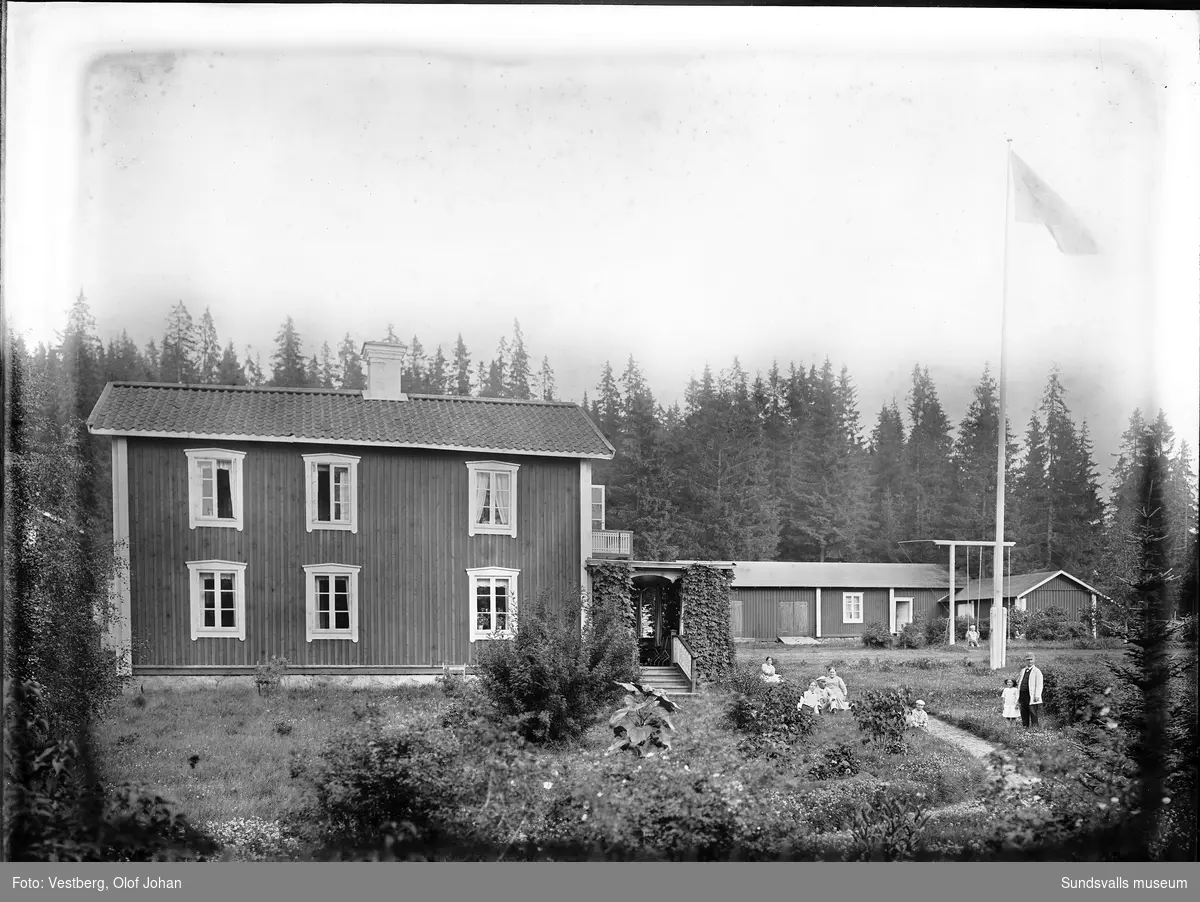 Gårdsbild i Skottsund med ett tvåvånings bostadshus, uthus och flaggstång. Vuxna och barn sitter och står i trädgården och en pojke gungar i en gungställning. Huset finns ännu (2024) kvar på adressen Skottsundsvägen 12.