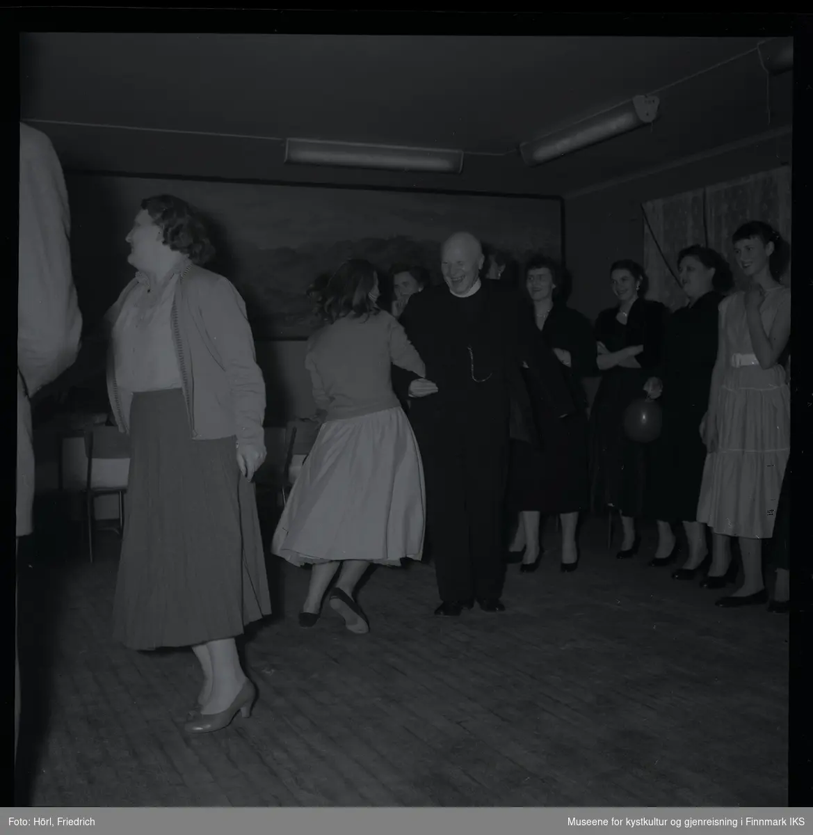 Den katolske menigheten i Hammerfest feirer juletrefest i 1957. Midt i bilde danser Pater Stockmann med en kvinne som har ryggen vendt til fotografen. På siden av dansegulvet er det flere kvinner som ser på sceneriet og er i godt humør.
