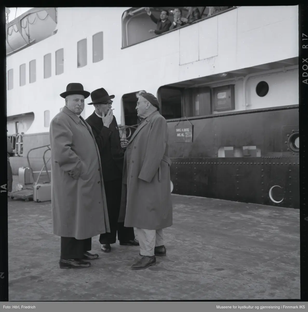 På kai i Harstad står tre menn og tar seg en røyk. I midten står Pater Spitz. En av de andre menn er Pater Rusche. I bakgrunnen ligger en skip til kais. Kofferter står ved landgangen og øverst i bildet ser man folk om bord på skipet der en av dem peker på noe.