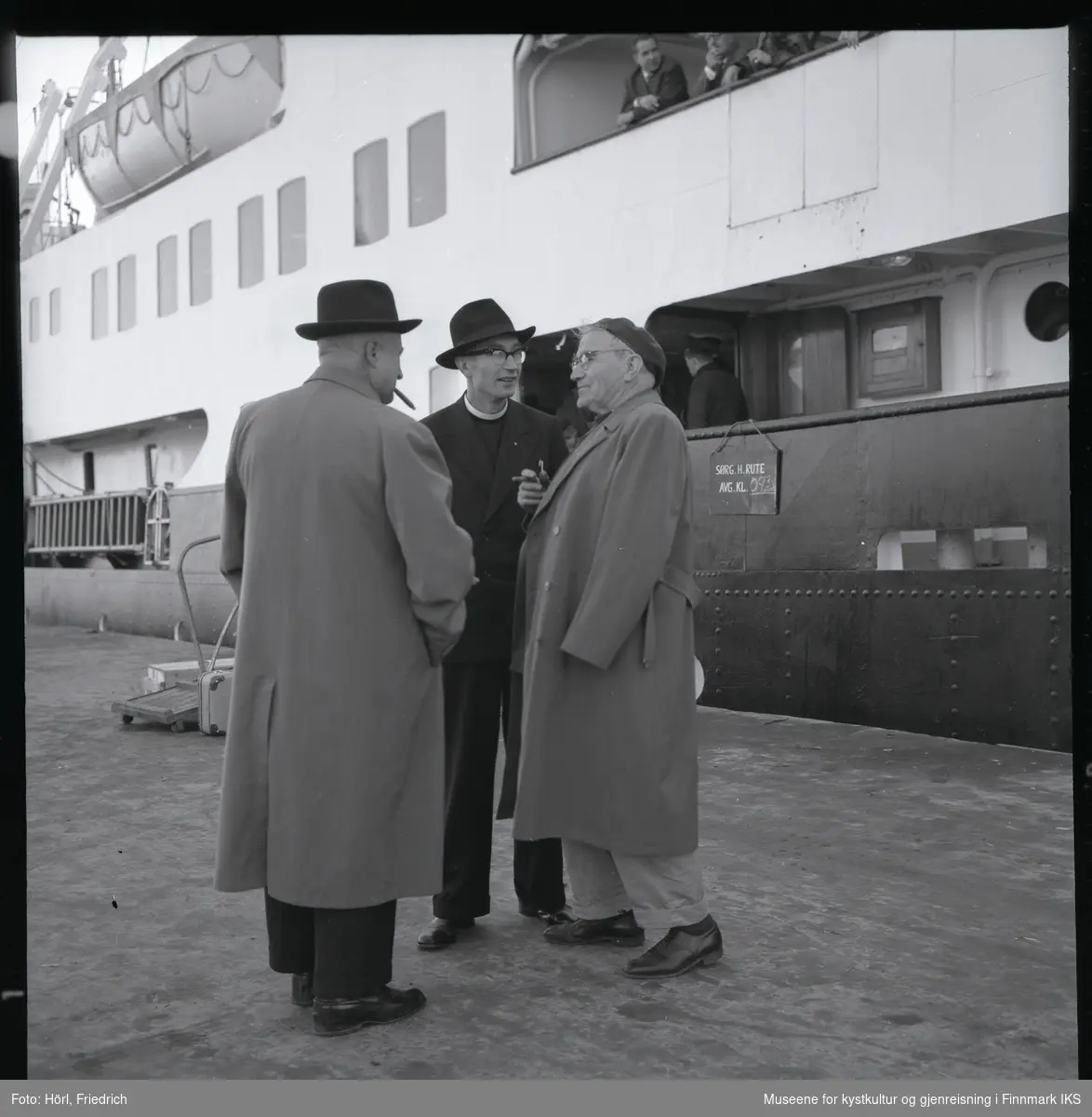 Tre men i samtale på kai i Harstad. To av dem tar røyk. I midten står Pater Spitz. En av de andre menn er Pater Rusche. I bakgrunnen ligger en skip til kais. Kofferter og transportgods står ved landgangen og øverst i bildet ser man folk om bord på skipet og en redningsbåt.