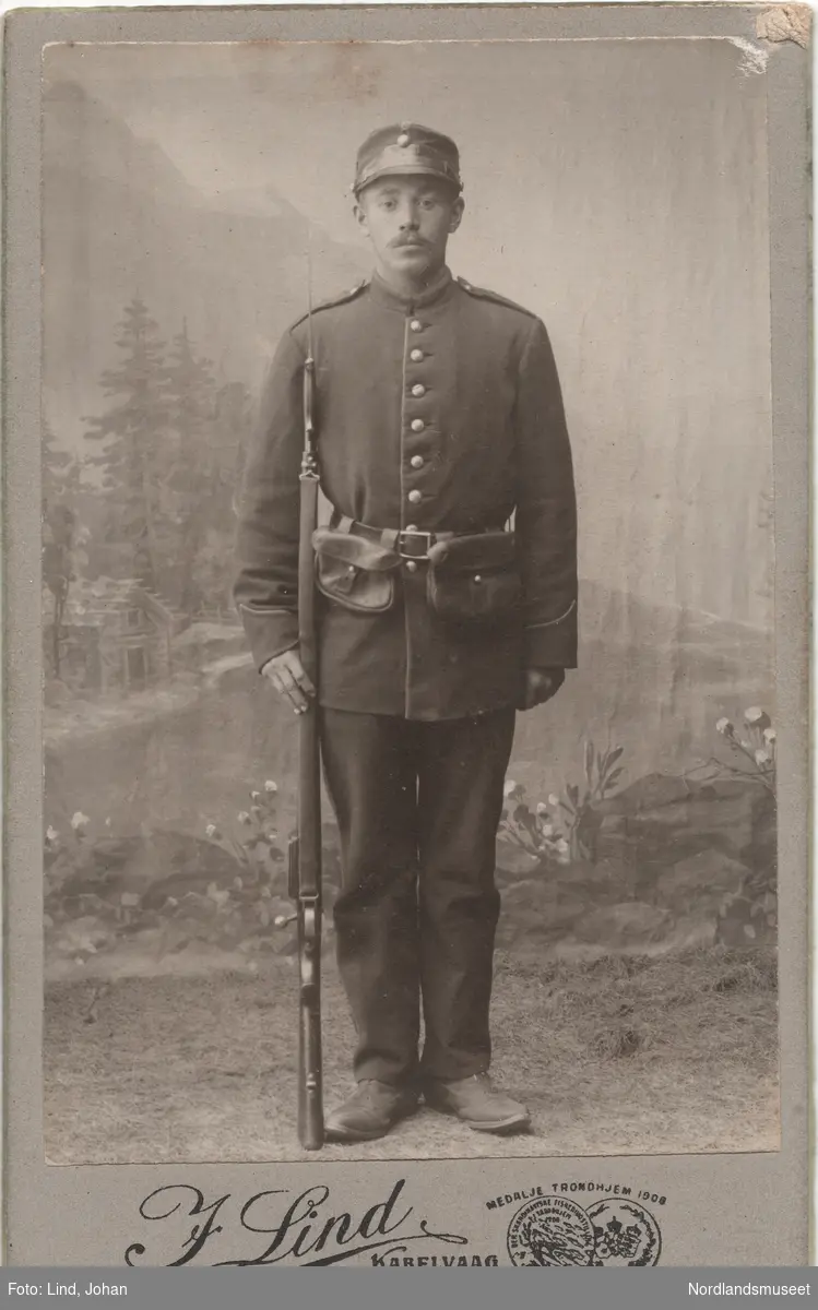 Portrett av en mann i uniform, stående med gevær på siden. Studiobilde foran malt lerret.