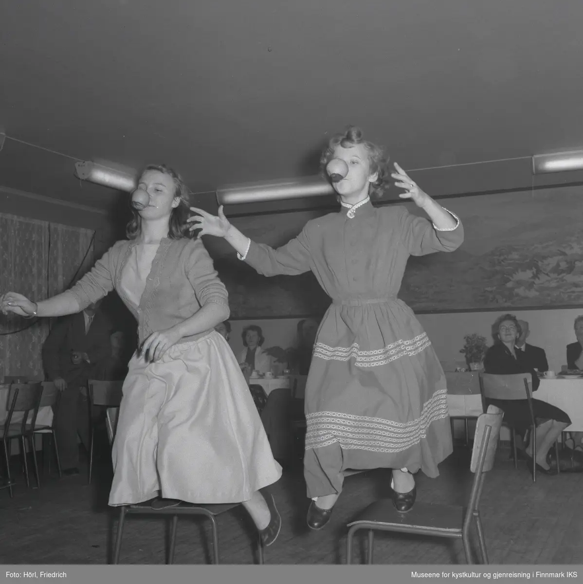 To unge damer har det moro på juletrefesten i den katolske menigheten i Hammerfest i 1957. De holder hver sin skje i munnen og balanserer epler på dem mens de går kapp over stoler. Dama til høyre er Lillian Trondsen. I bakgrunnen ser man flere personer som sitter ved bordene og ser på.