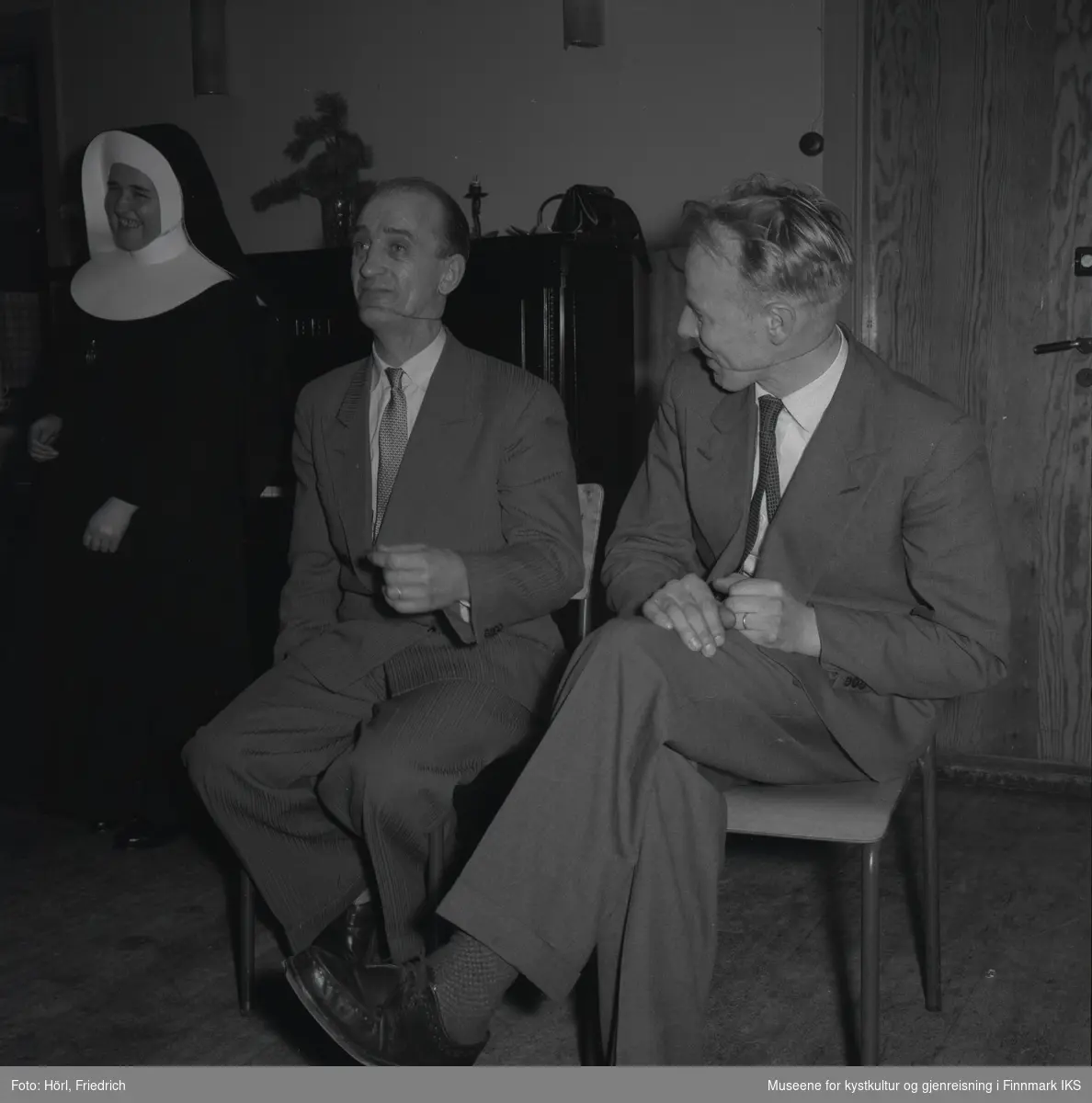 Theodor Trondsen til venstre og Per Eriksen til høyre, har det moro på juletrefest i den katolske menigheten i Hammerfest i 1957. De sitter på stoler ved siden av hverandre og har gummistrikk rundt hodene og tvers over ansiktene. Til venstre for mennene står en nonne som brister ut i latter. I bakgrunnen står en piano.