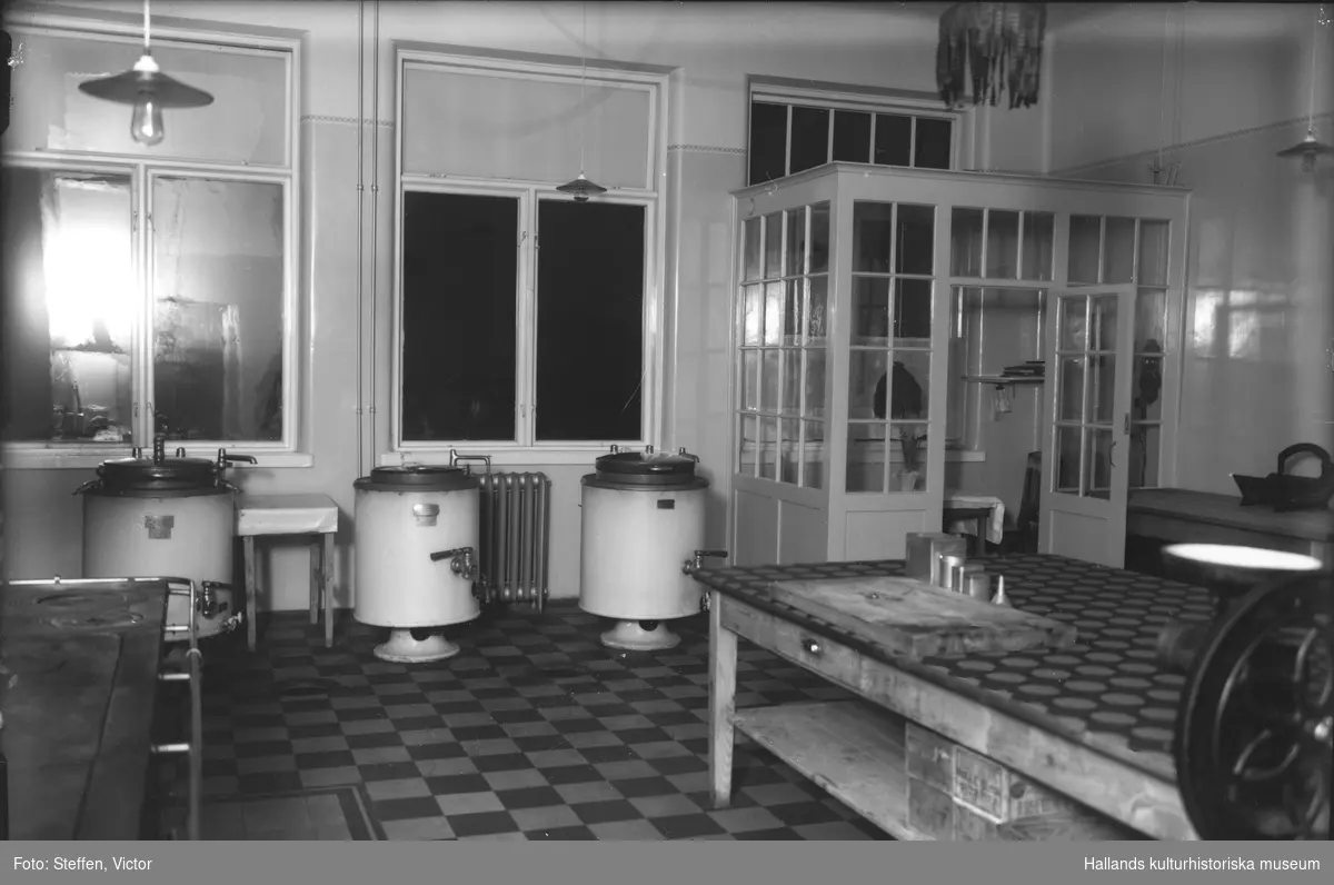 Interiör av köket på Fagereds sanatorium, 2 fotografier. Köket har vedspis och mitt i rummet ett stort bord med en skivapparat (skinka, bröd etc), en skärbräda, olika bleckburkar, en tratt och vid ett hörn är en korvstoppningsapparat fästad. Vid fönstren står tre cylinderformade maskiner med olika kranar, liknande centrifuger, kanske en form av varmvattenberedare. I hörnet ett inglasad litet personalutrymme med telefon.