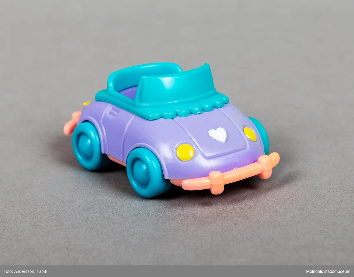 Lila leksaksbil i plast med gröna hjul och rosa kofångare. Baktill på bilen finns en rosa rosett.