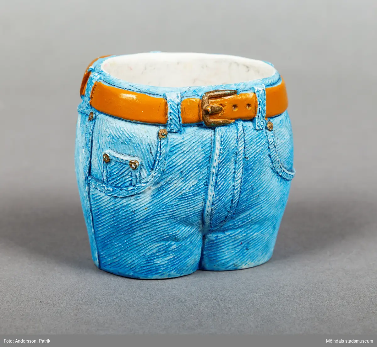 Pennställ i porslin, utformat som linningen på ett par jeans med ett bälte i midjan. Jeansen är ljusblå och bältet är ljusbrunt. Framtill har byxorna två fickor och en gylf. Baktill finns också två fickor. Ur en av bakfickorna sticker det upp en kam.
