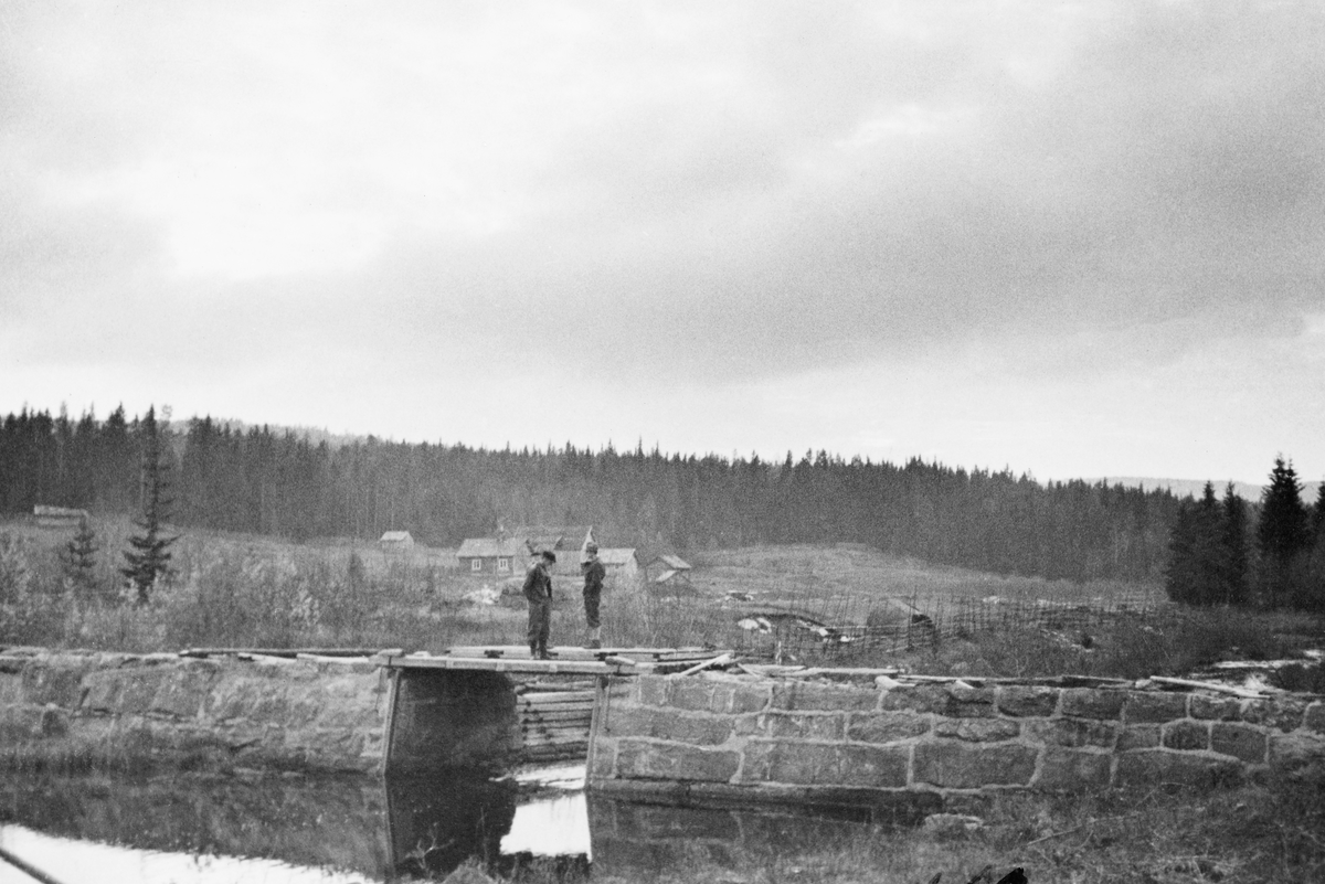 Gjeddevassdammen i Sæteråa i Sør-Odal kommune i Hedmark høsten 1933. Bildet er tatt i medstrøms retning. Her var damarmene murt av bruddstein. Gjeddevassdammen hadde ett løp med «golving» eller «flakning» i botnen, og med steinfylte tømmerkister som skådammer i forlengelsen av damløpet. Dambrua var av tre. På den lå det fire luker som, i fløtingssesongen, kunne heves og senkes for å tilpasse vannstanden i den nedenforliggende delen av vassdraget fløternes behov. Sæteråa kommer fra fire små sjøer i grensetraktene mot Sverige - Sæterlisjøen, Skårillen, Grønsjøen og Mjøgsjøen. Også ved utløpene av alle disse fire sjøene var det dammer som gjorde det mulig å bruke dem som reservoarer for fløtinga. Det fantes også en fløtingsdam ved Sætersaga, i den nedre delen av det snaut to mil lange vassdraget, som renner inn i Glomma ved Disenå. Da dette bildet ble tatt sto to menn - antakelig fløtingsfunksjonærer på befaring - på dambrua. Det året dette fotografiet ble tatt var det innmeldt 22 575 tømmerstokker til fløting i Sæteråa.