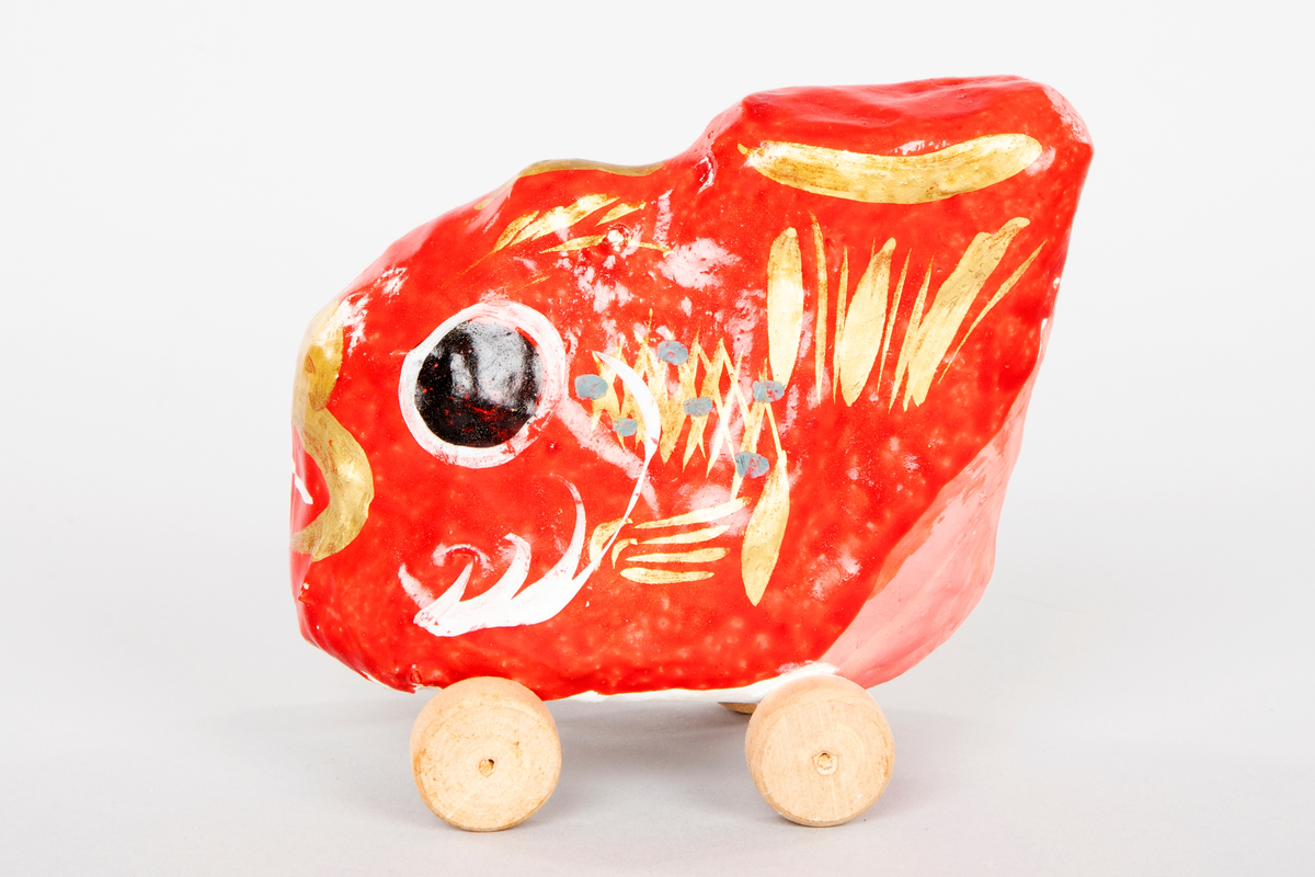 Fisk på hjul i pappmasjé og gips. Figuren er malt blank rød med store svarte øyne. Fisken har gullfarget munn, rygg og finne, noen blå prikker på kroppen og er matt oransje på undersiden. Hjulene er laget i tre.