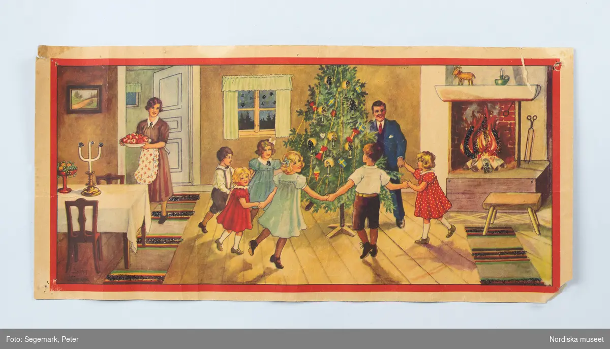 Plansch julfirande, utförd av Jenny Nyström. En man och några barn dansar kring julgranen. En kvinna bär ett fat med röda äpplen. I den öppna spisen är en brasa tänd.