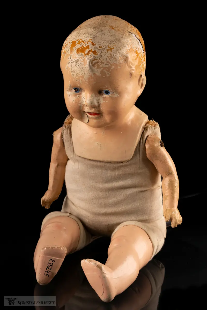 Dukke med hode, armer og ben av malt pappmasje, og kropp av grov bomullsstoff stoppet med halm. En fløyte som simulerer barnegråt når dukken snus er integrert i kroppen.