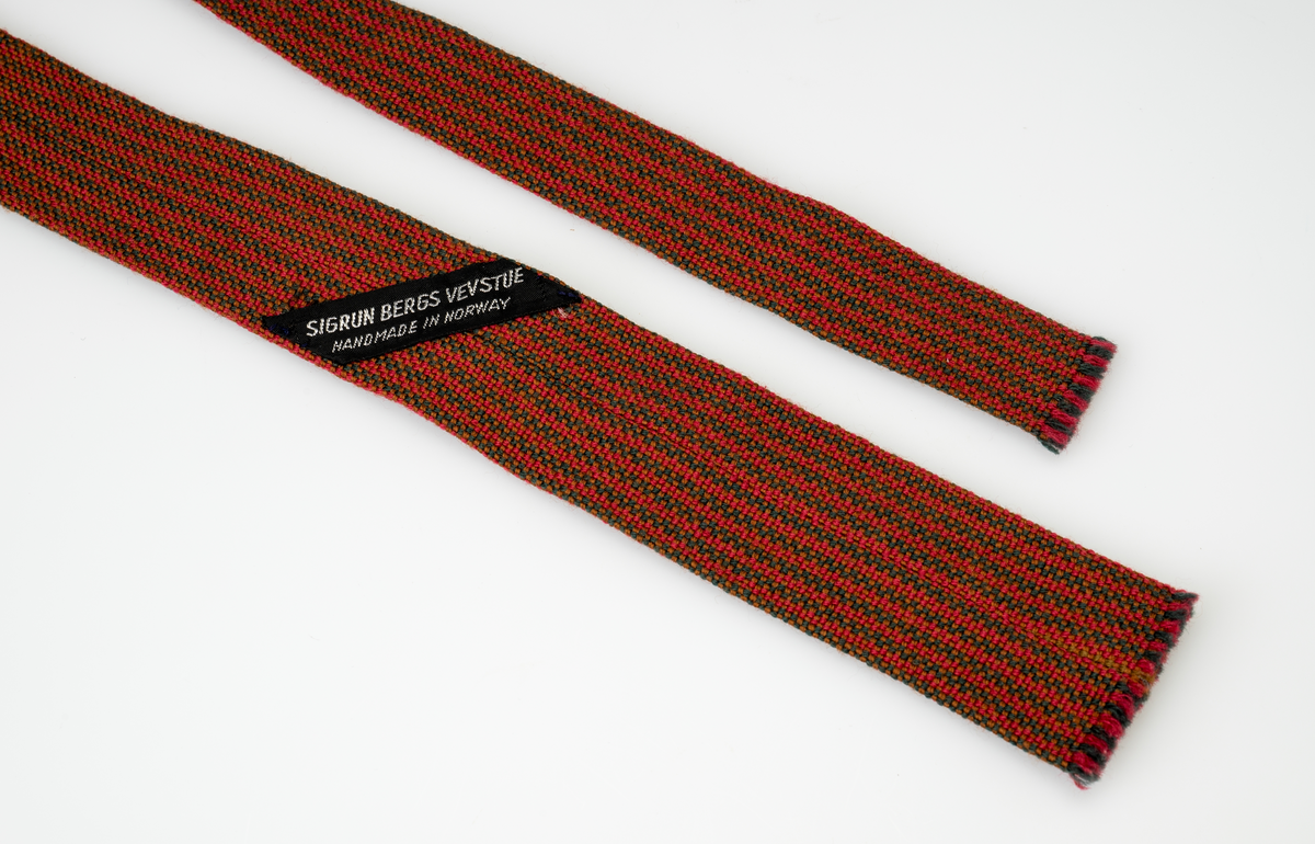Et smalt og langt slips håndvevd av ullgarn i fargene rødt, grønt og brunt. Fargene utgjør et stripe- og rutemønster. Det vevde stoffet er brettet slik at det er dobbelt og sydd sammen med maskinsøm på vrangen. I hver ende av slipset er det korte frynser (0,2-0,3cm). Slipset er smalere i den ene enden enn den andre.