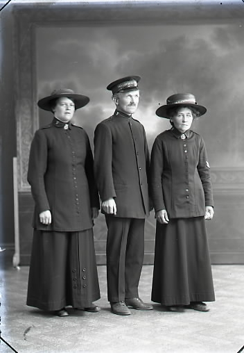 Ateljébild. Tre frälsningssoldater, två kvinnor och en man, stående i helfigur. Samtliga iförda uniform med märken och hattar. Beställare: John Persson.