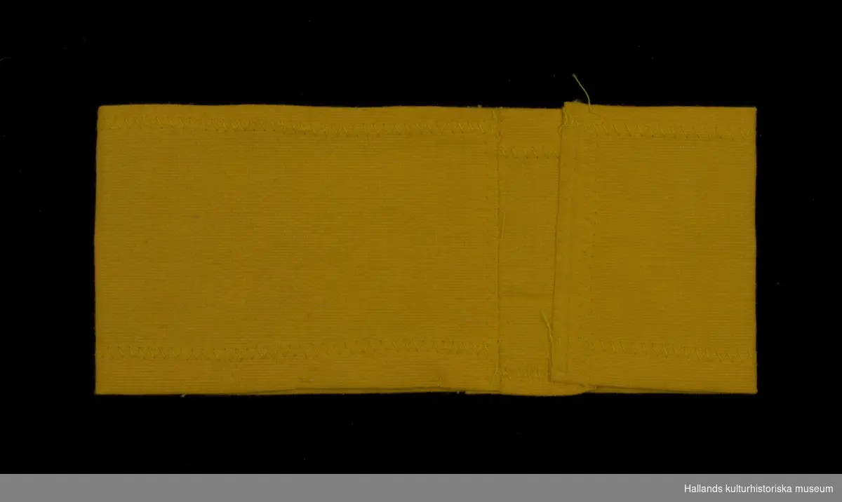 Armbindel av gult bomullstyg. Påsydd antikärnkraftssymbol: gul botten med röd skrattande sol samt svart text: "ATOMKRAFT? NEJ TACK".