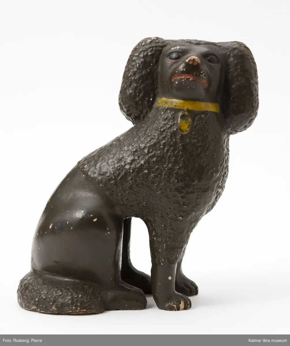 KLM 12118. Figurin. Tillverkad av lergods. Figurin i form av en sittande hund, pudel, brunfärgad, med gult halsband, röd mun och nos, ögon i svart och rött. Stämpel: Augusta, JB.  Par med KLM 12119.