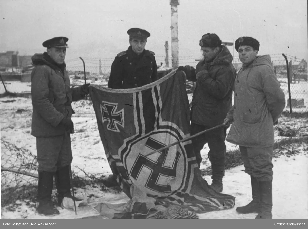 Gruppebilde av 4 sovjetiske soldater som viser frem et nazi-tysk flagg i ruinene av Kirkenes by, høsten 1944.  