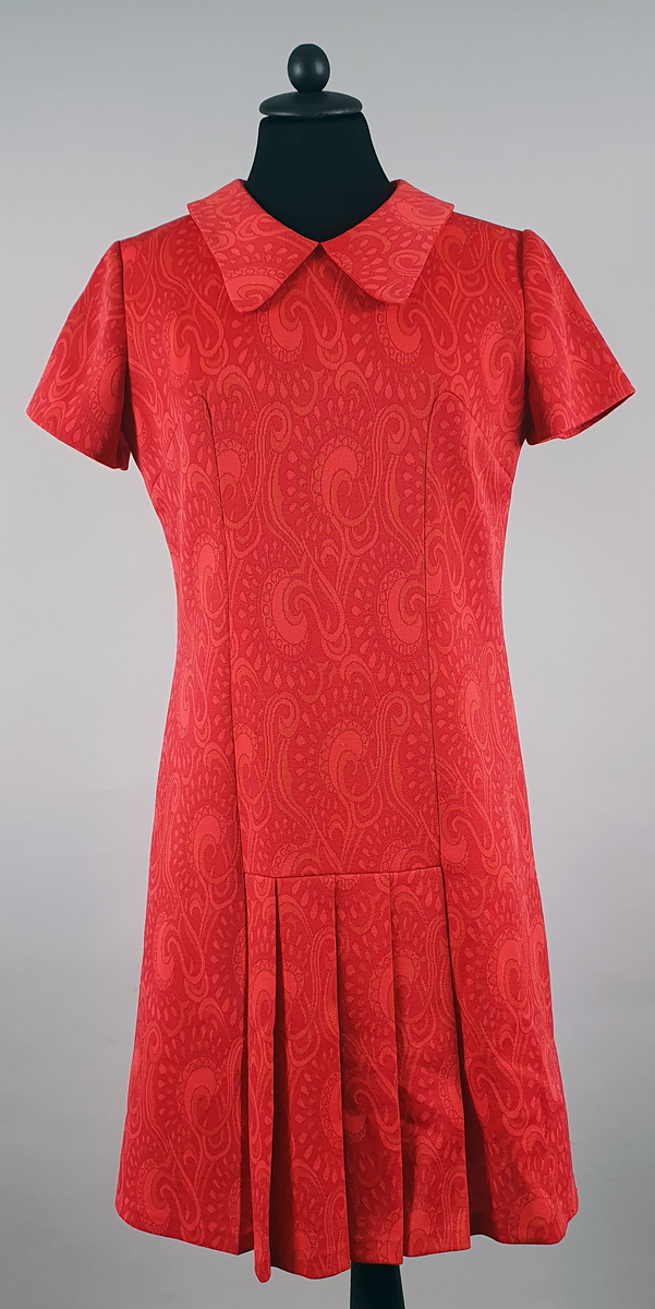 Rød kjole med orientalske mønster. Rett fasong med korte ermer og plisséfolder midt foran. Skjortekrage og glidelås i ryggen.