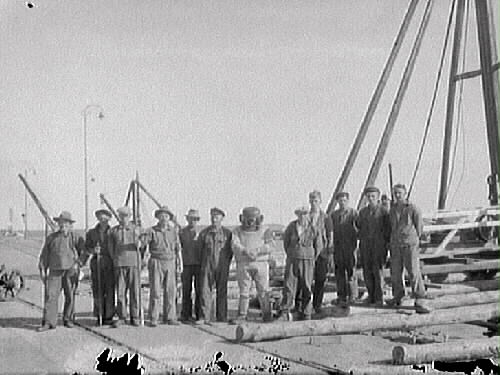 Dykare i full mundering står med en samling män på kajen i Varbergs hamn. De till vänster ser ut att hålla i spett och i förgrunden ligger några stockar.