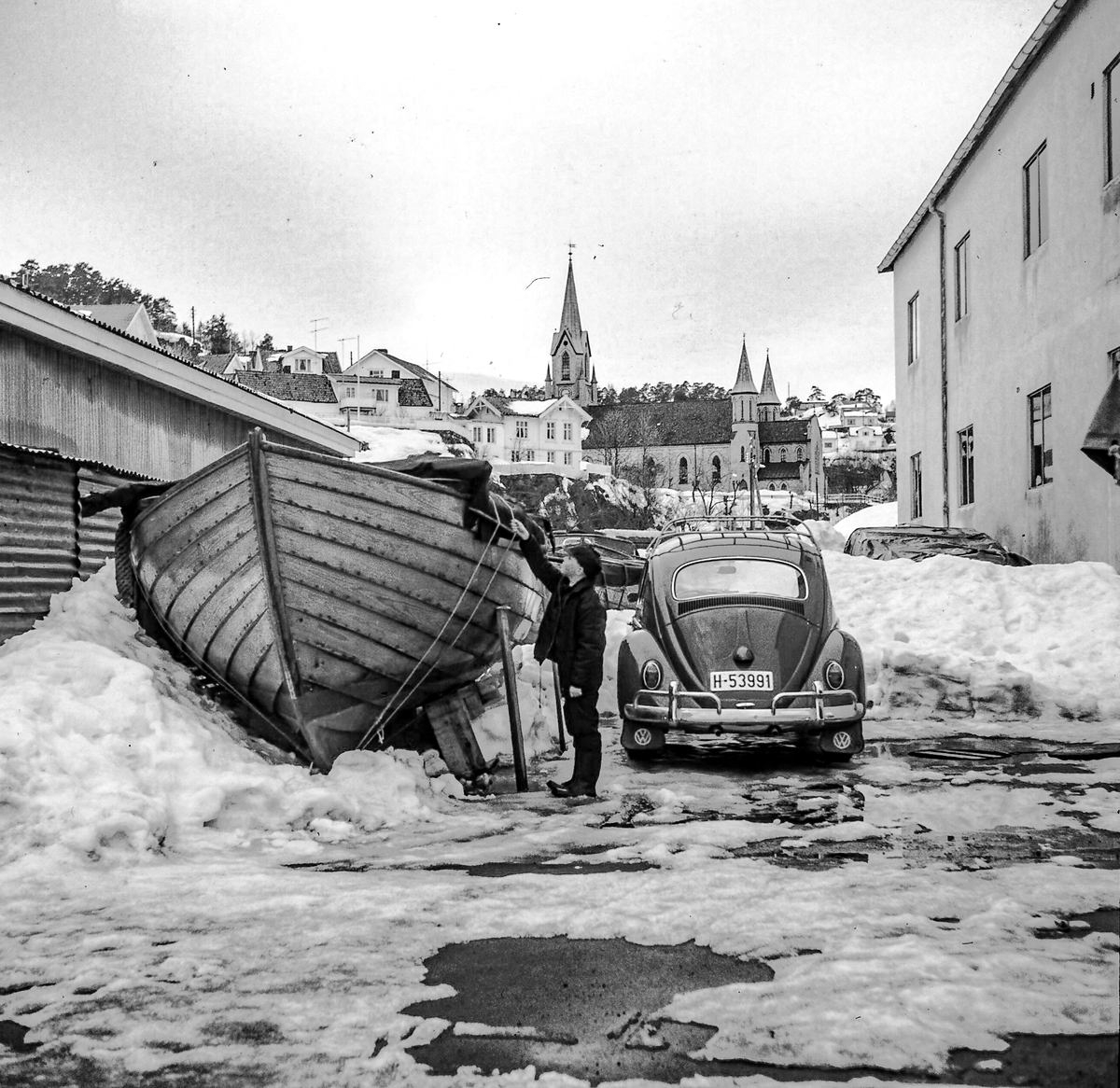 Mann sjekker koggen i vinterlagring ved felleskjøpet. 1960-70. Kragerø kirke bak.