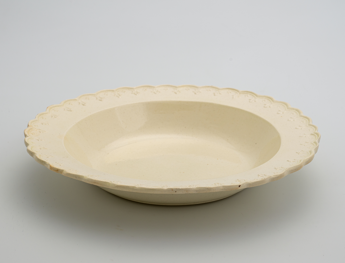 En dyp tallerken av glasert steintøy (creamware). Den er hvitgul på farge (ensfarget). Langs kanten er det tungekant. Innenfor tungekanten er det støpt dekor i form av en hjertebord med stiliserte blomster.