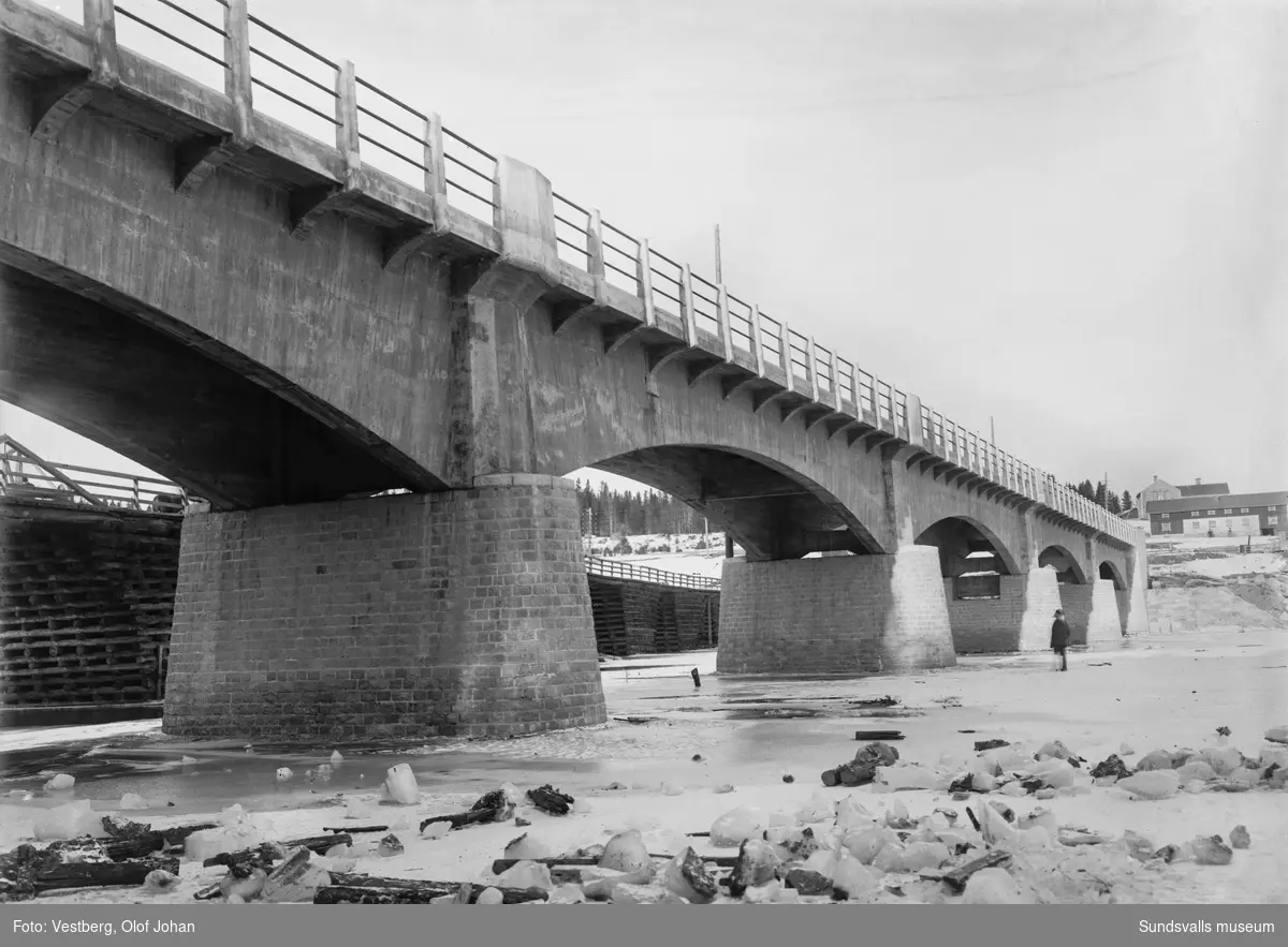 Vintervy norrut vid den nybyggda betongbron i Njurundabommen, bakom syns den ännu ej rivna gamla träbron. En man står på isen och tittar på bron. Gården som syns norr om bron finns ännu (2024) kvar.