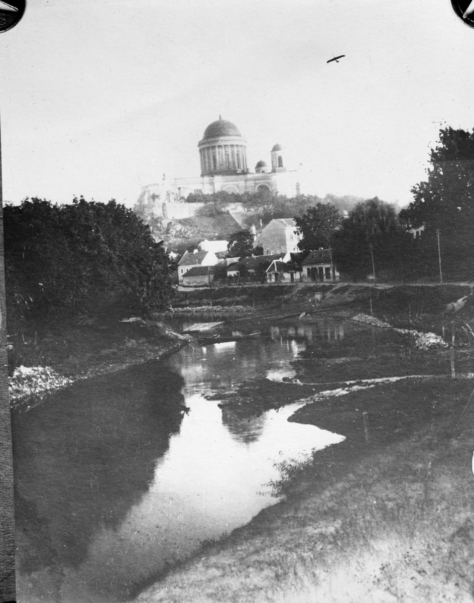 Flygplan Etrich Taube i luften över basilikan (kyrkan) i Eztergom i Österrike-Ungern, omkring 1910-1915. Vattendrag i förgrunden. Reprofotografi.