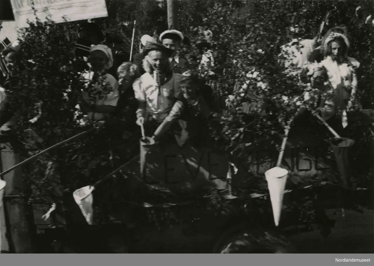 En lastebil pyntet med bjørkekvister. I lasterommet står det unge kvinner i pene klær. Bilde tatt i Bodø under feiring av freden etter okkupasjonen 1945.