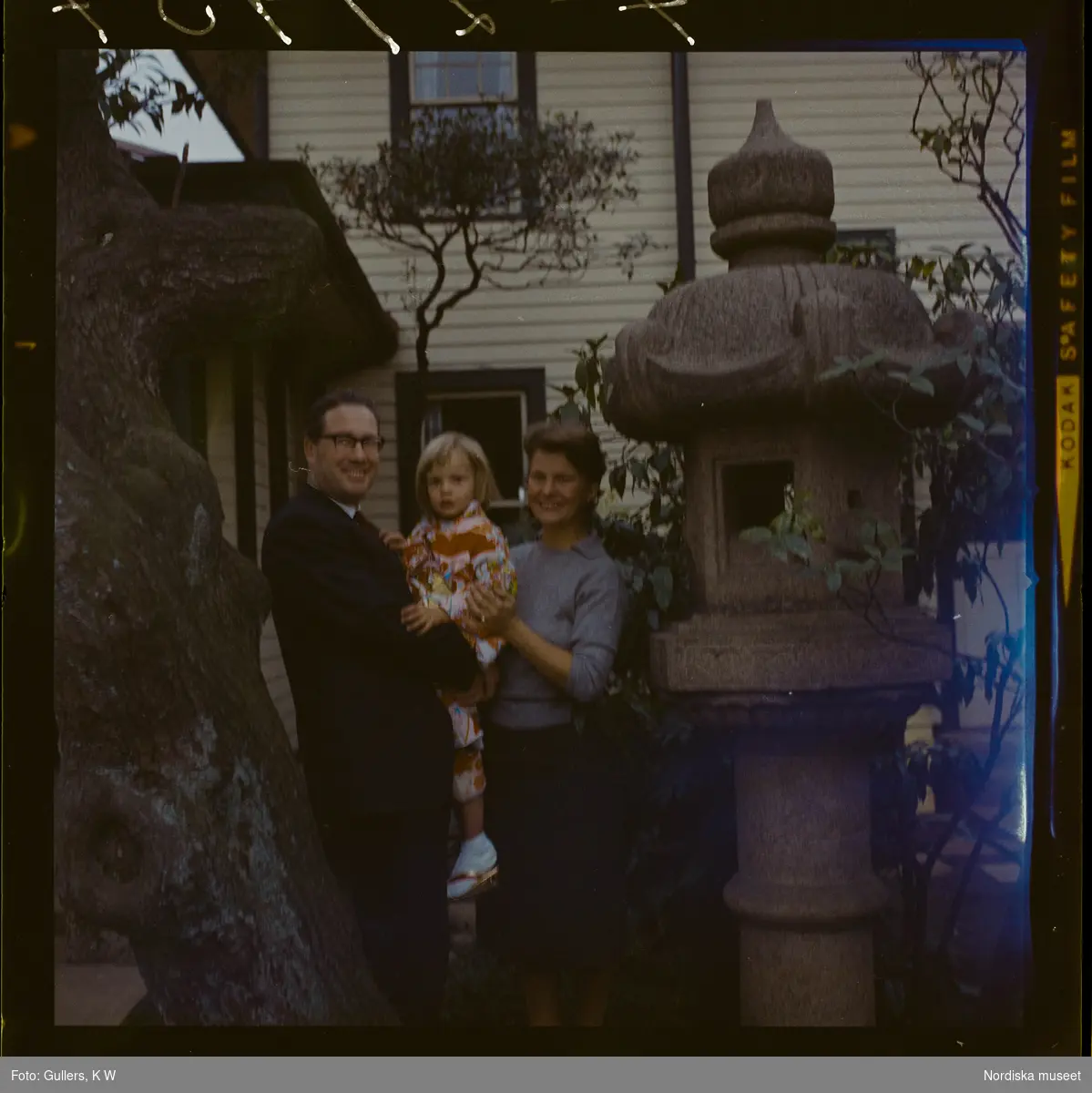 585/8 Facit Tokyo Daberg. En familj står i en trädgård vid en japansk lykta av sten. Barnet är klädd i kimono.