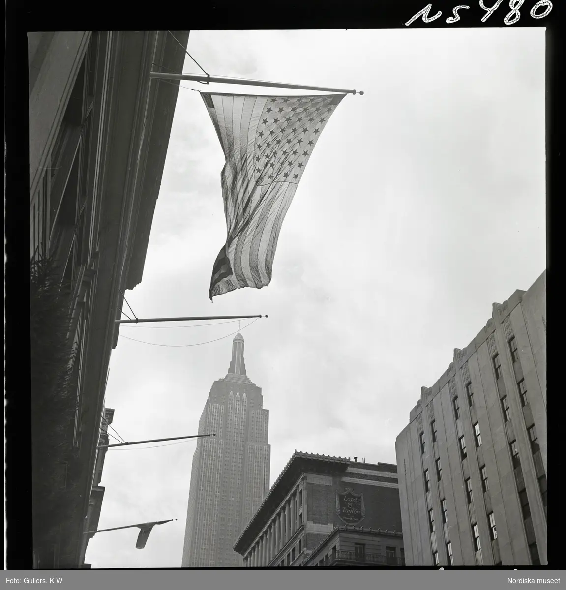 1690 New York allmänt (N.Y. Herald Tribune). Gatubild. Amerikansk flagga hänger från fasad, Empire state buildning i bakgrunden.