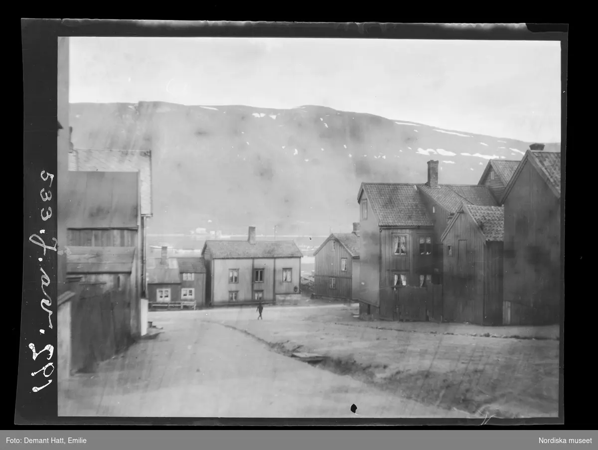Stadsbild från Tromsø i Norge. Låga trähus kantar gatorna, i bakgrunden anas ett fjäll. En ensam människa står på en gräsbevuxen yta. Bilden ingår i en serie fotografier tagna av Emilie Demant Hatt i Sapmi mellan åren 1907 och 1916.