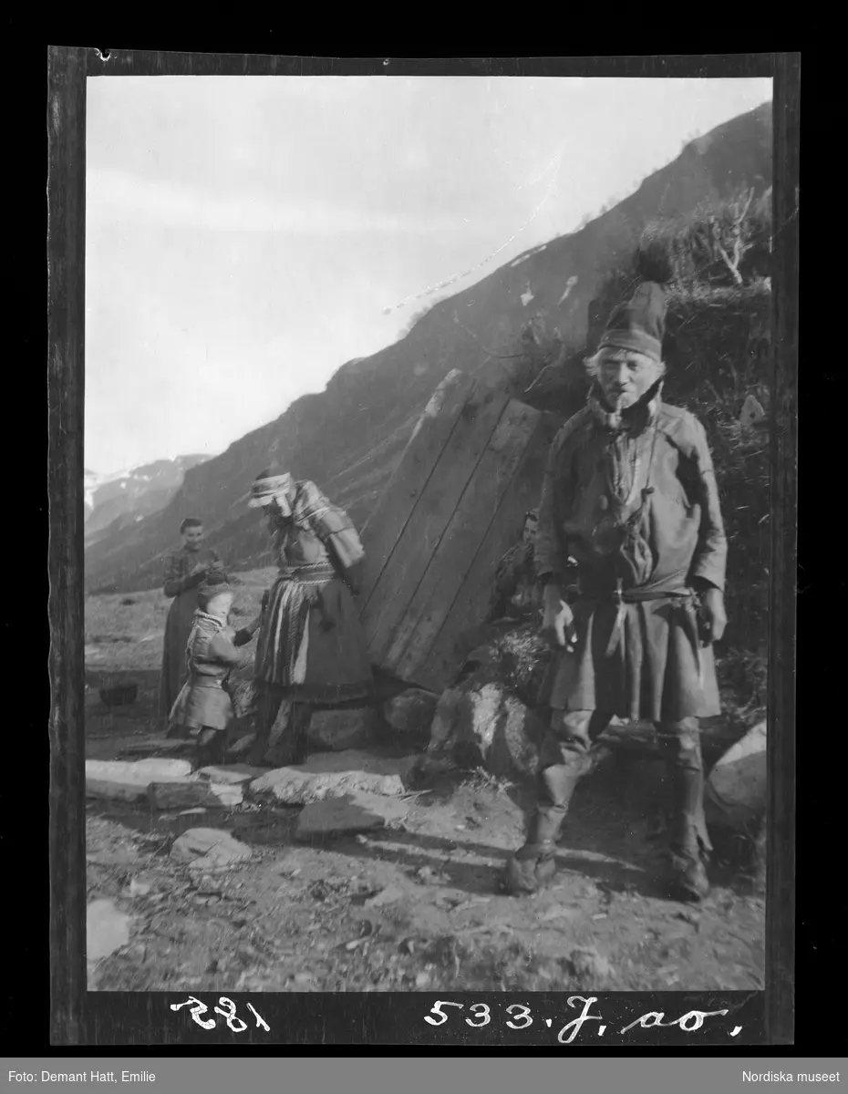 Framför en kåta står en man, Hoalsta-Lasse. bakom ses kvinnor och barn. I bakgrunden fjäll.  Bilden ingår i en serie fotografier tagna av Emilie Demant Hatt i Sapmi mellan åren 1907 och 1916.