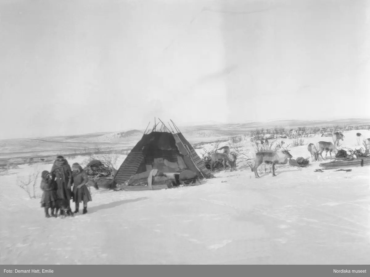 Tre flickor står i snön framför en kåta vid en lägerplats under vårflyttningen från Närvä. Där finns också ett antal renar. Bilden ingår i en serie fotografier tagna av Emilie Demant Hatt i Sapmi mellan åren 1907 och 1916.