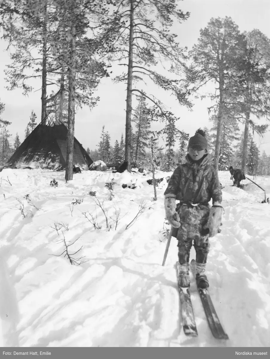 Pojke, Andaras (Nilsa), på skidor i snön, troligen under höstflyttningen vid Laimo. I bakgrunden ses en kåta och en person som gräver snö (till kaffekokning). Bilden ingår i en serie fotografier tagna av Emilie Demant Hatt i Sapmi mellan åren 1907 och 1916.