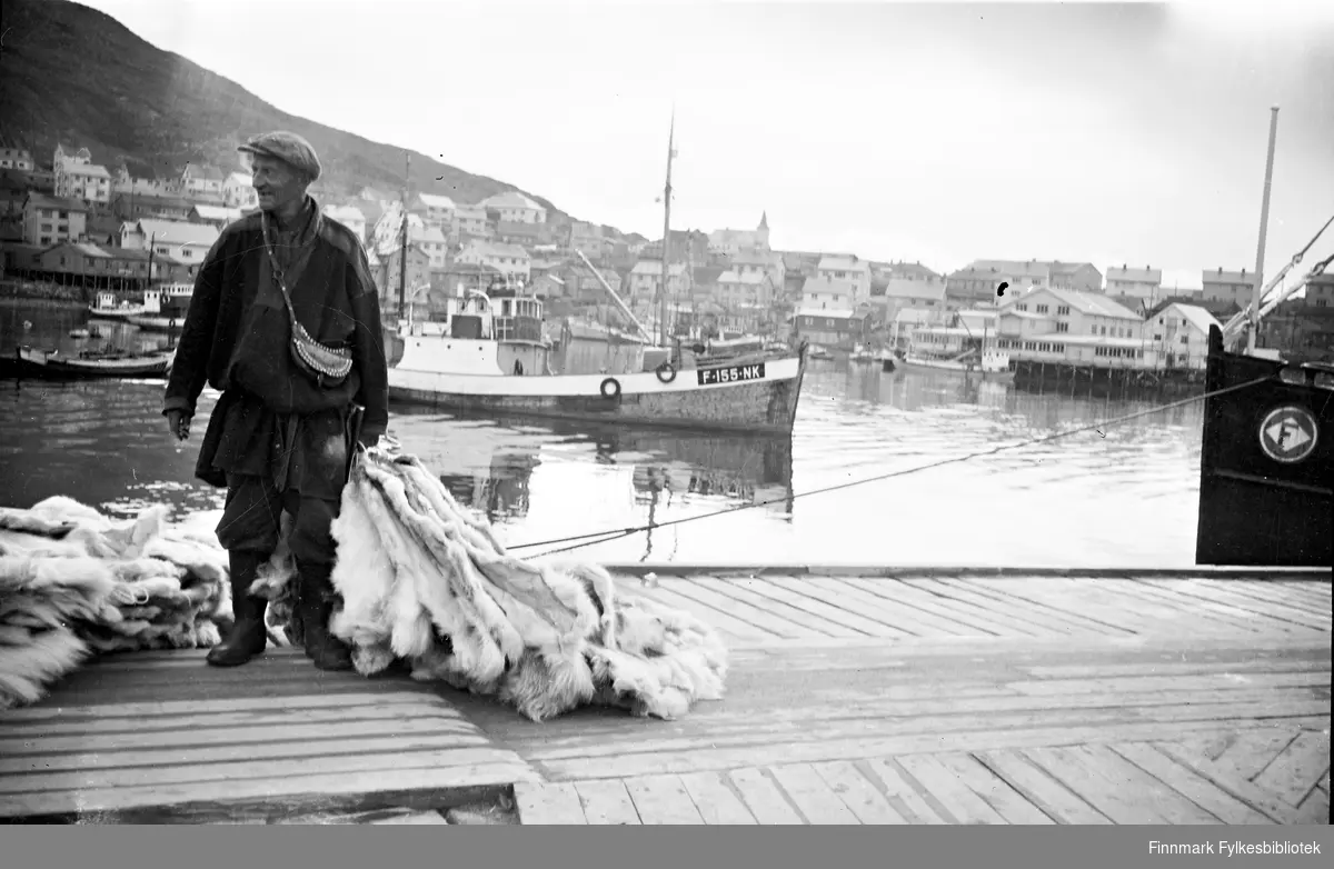 Honningsvåg havn ca.1955. En samisk mann selger reinskinn på kaia. Flere fiskebåter og hus i bakgrunnen. Vi ser også kirka som var den eneste bygningen som stå igjen etter krigen.