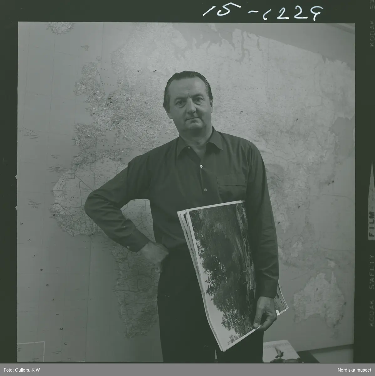Fotograf K W Gullers står framför en väggkarta med ett antal fotoprintar under armen.