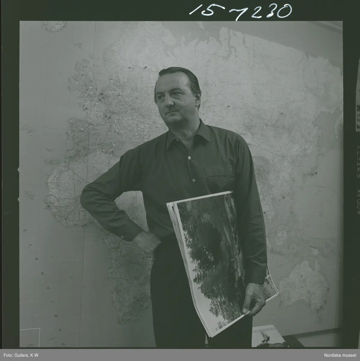 Fotograf K W Gullers står framför en väggkarta med ett antal fotoprintar under armen.