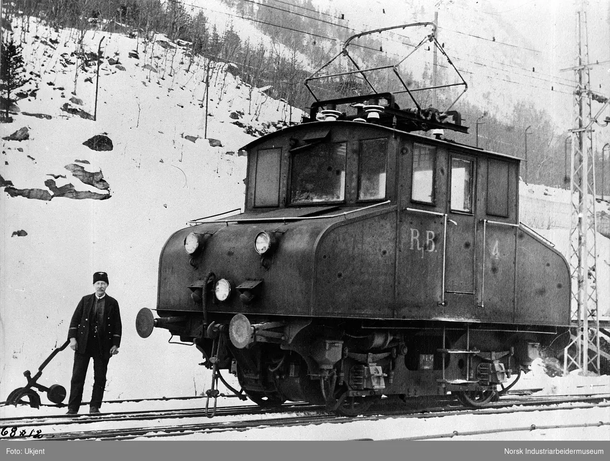 Mann kledd i jernbaneuniform stående foran elektrisk lokomotiv RjB 4. Sporveksler ved jernbanesporet.