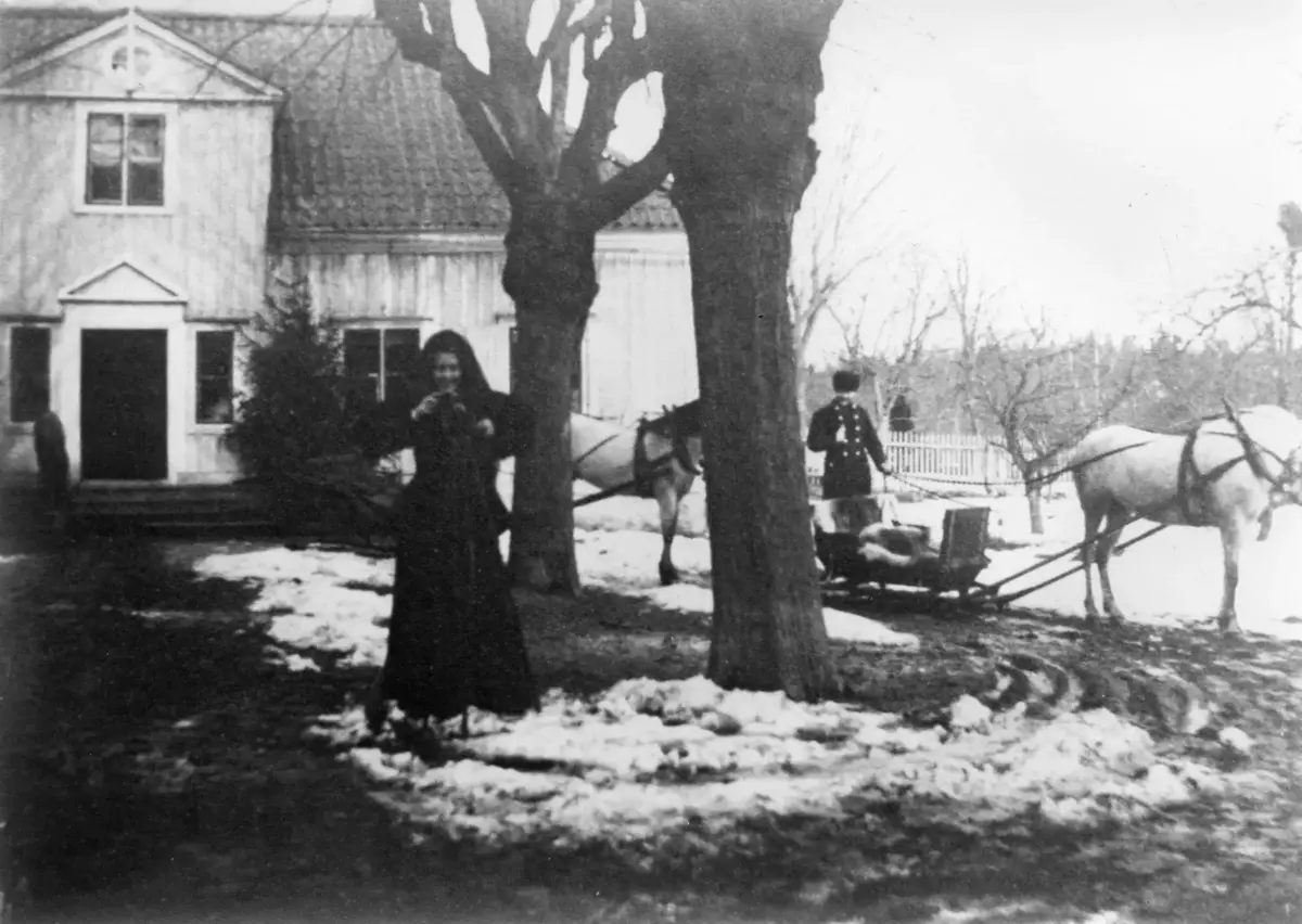 På gårdstunet gör en kvinna sig beredd att ge sig ut på slädparti. Två hästekipage i bakgrunden, samt en uniformsklädd kusk